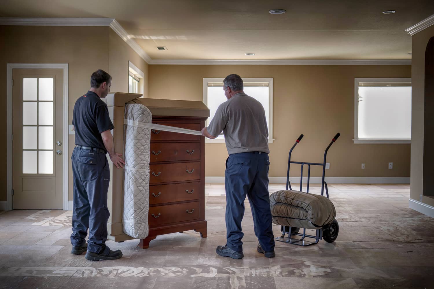 Two movers wrapping a wooden dresser in quilted blankets and stretch wrap in an emptied room