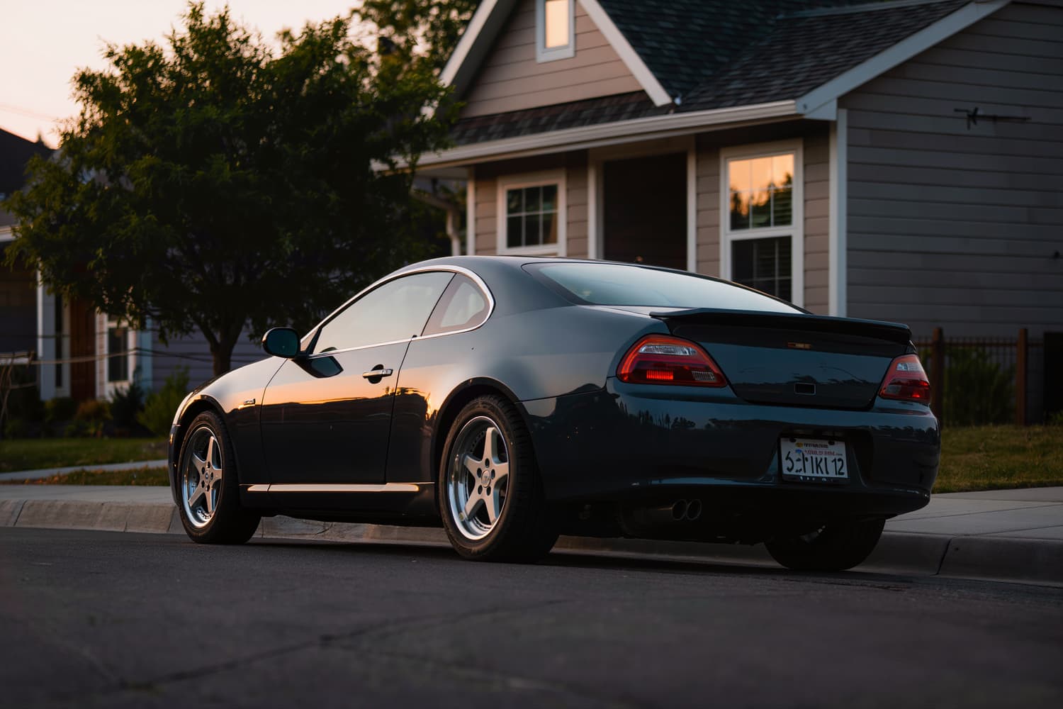 Dark coupe at the kerb of a residential street at sunset, low rear three-quarter view