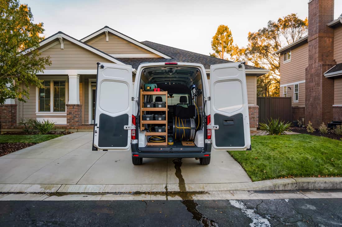 Unmarked white service van backed into a driveway with rear doors open on fittings and a hose reel