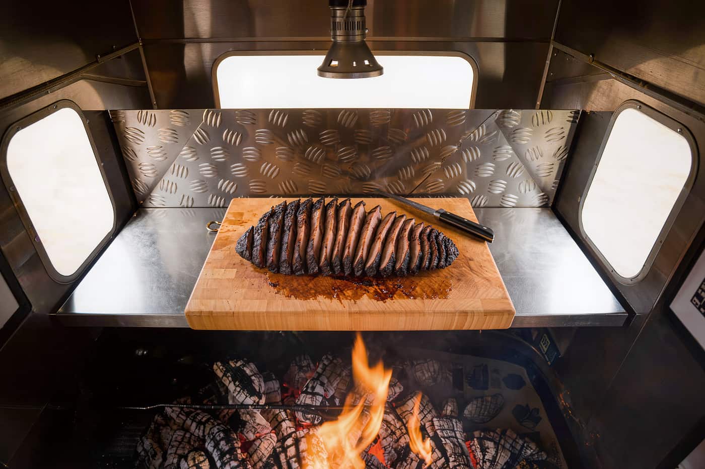 Sliced brisket fanned across a wooden board on a stainless prep counter inside a barbecue trailer.