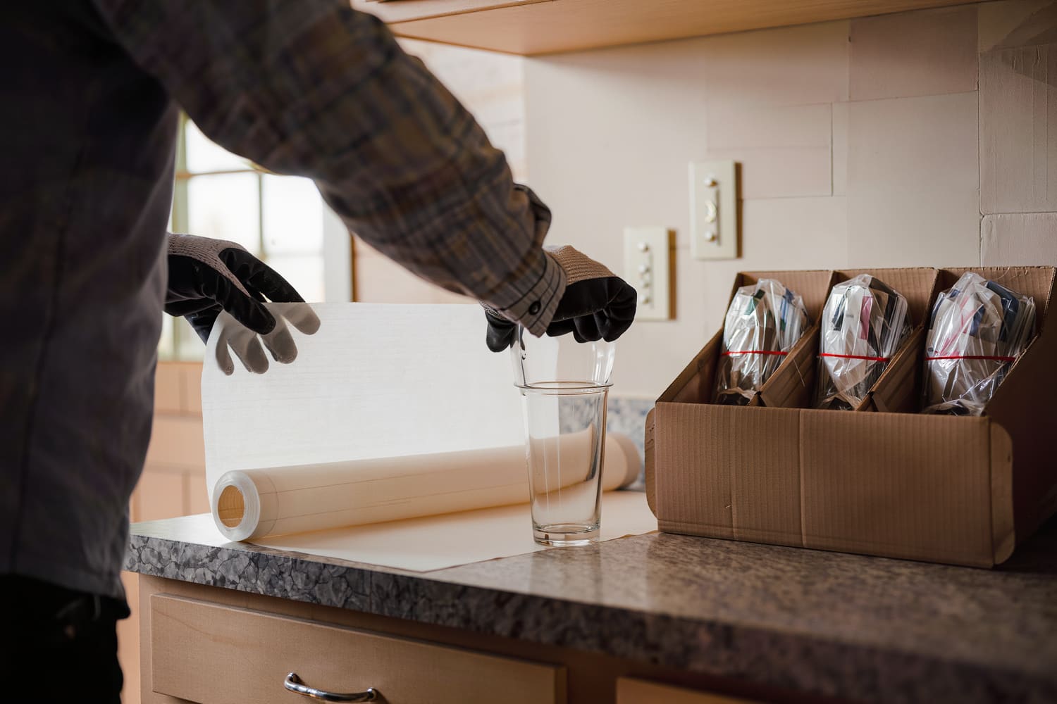Gloved hands rolling a drinking glass into packing paper at a kitchen counter.
