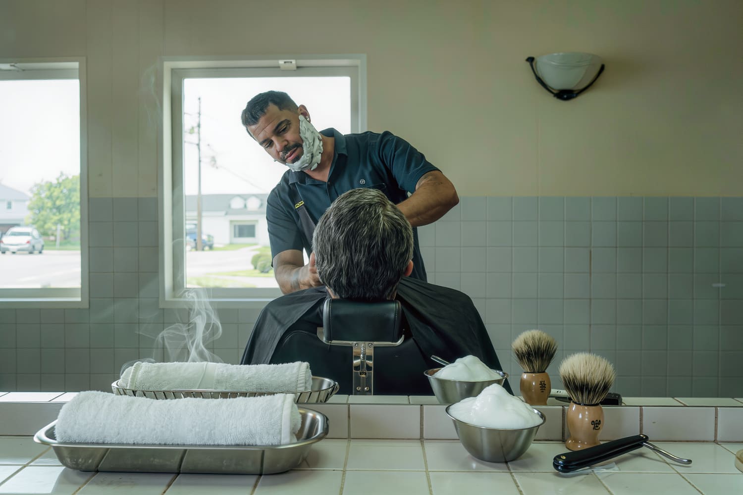 Barber from behind at a reclined chair, steam rising off a rolled towel in a tray