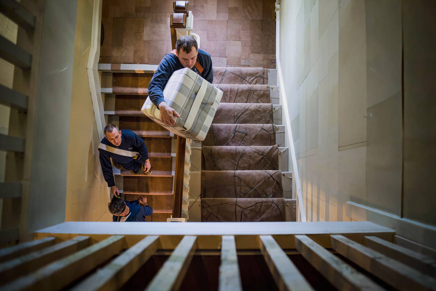 Movers carrying a blanket-wrapped carton up a carpeted staircase protected with a runner