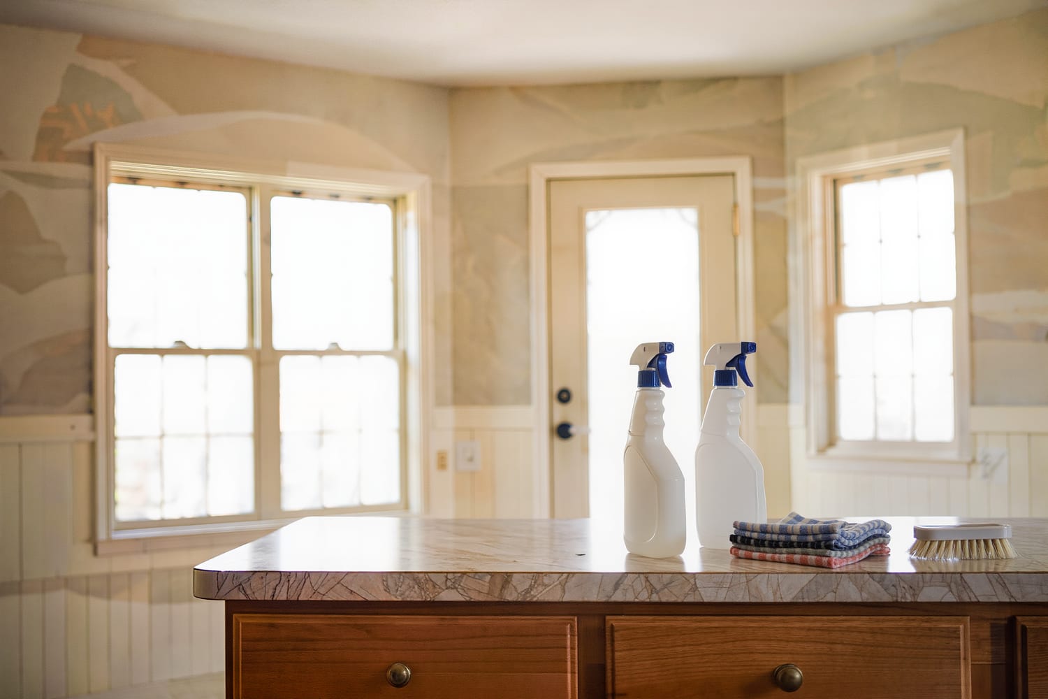 Unlabelled spray bottles, folded microfibre cloths and a caddy staged on a kitchen counter