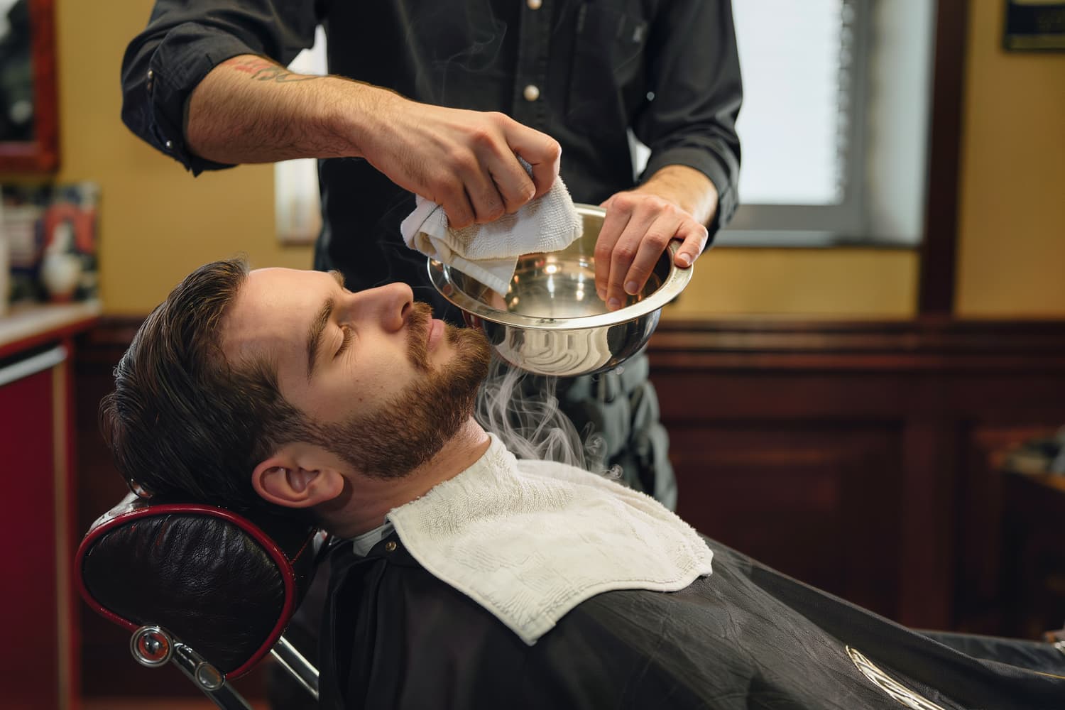 Barber's hands lifting a steaming folded towel from a steel bowl at the shave station