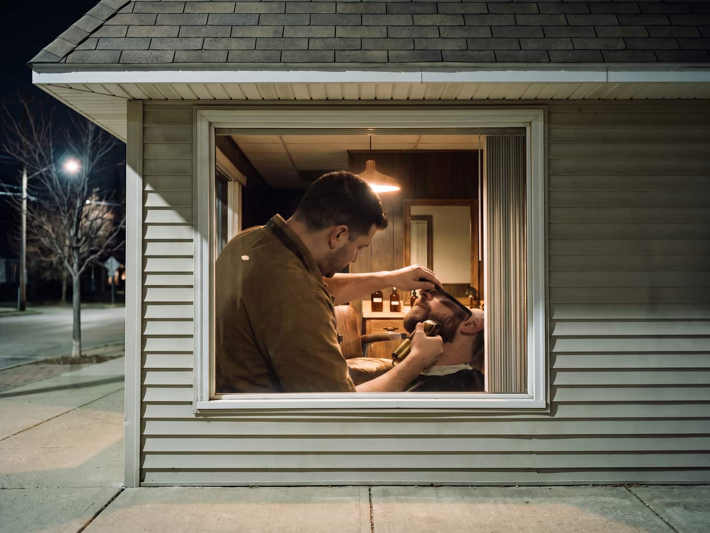 A barber trimming a client's beard, seen through the shop window at night