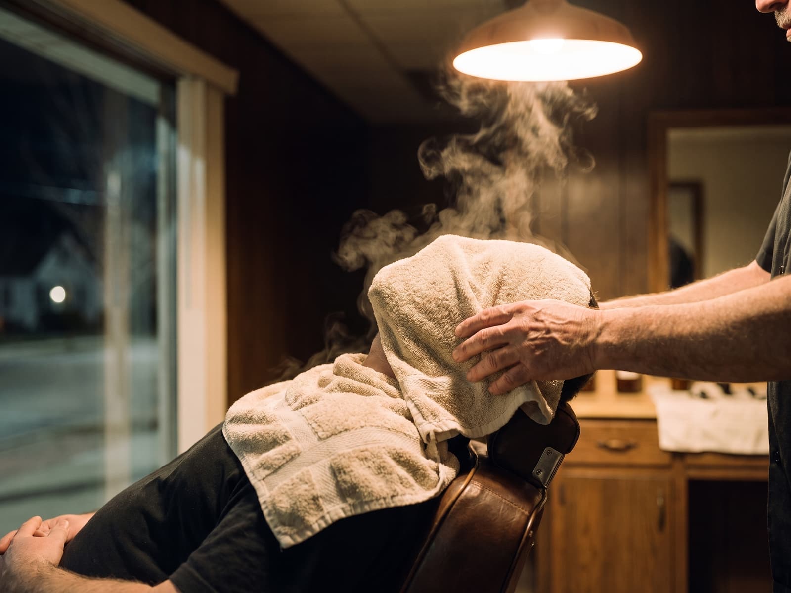 Hot towel being lifted from a reclined client's jaw, steam rising