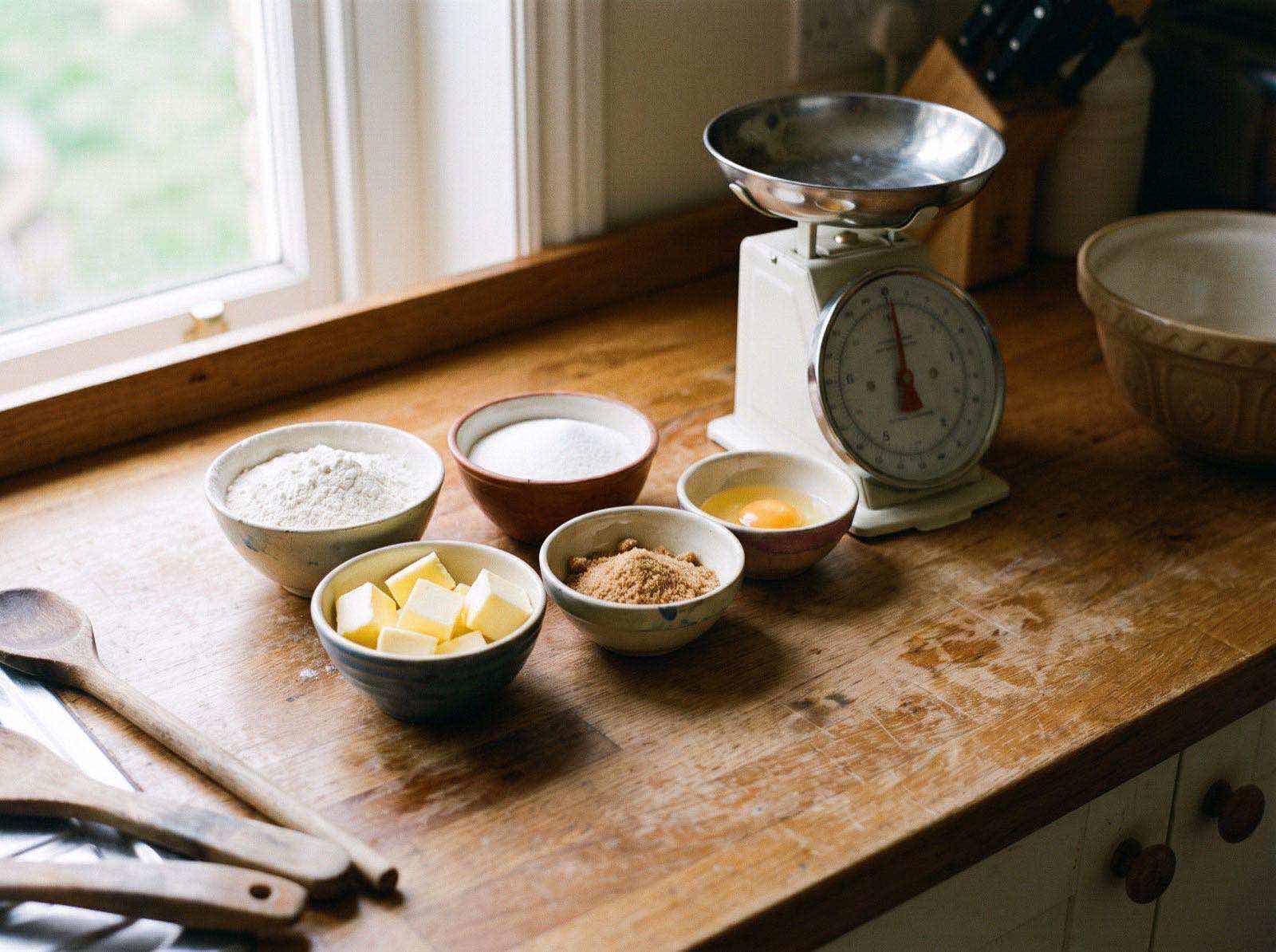 Flour, sugar, cubed butter, brown sugar and a cracked egg measured into small bowls beside a kitchen scale