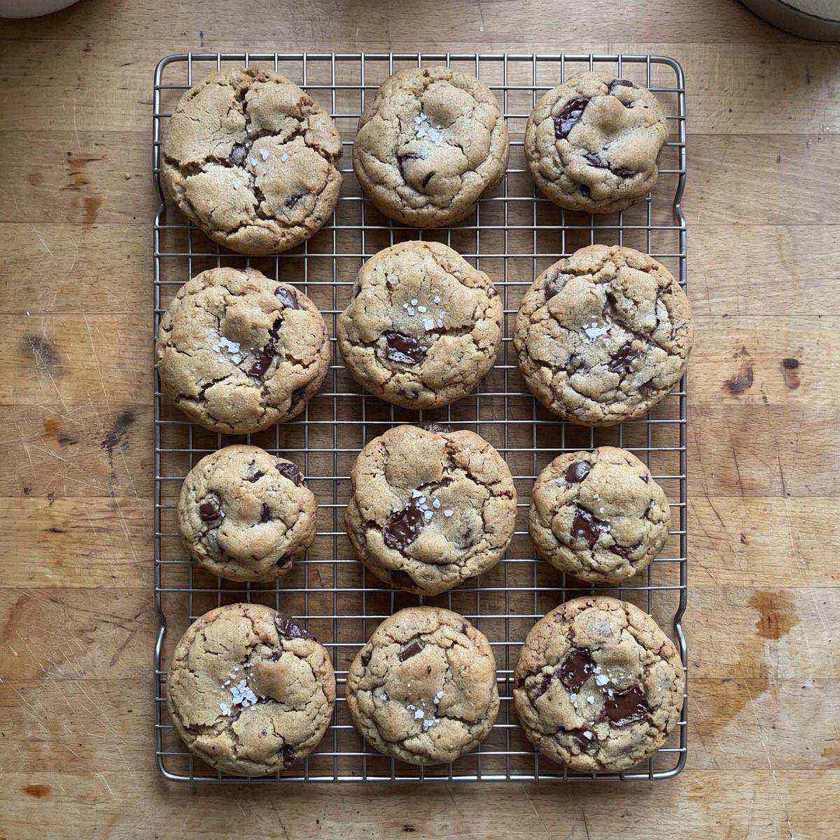 A dozen chocolate chip cookies with cracked tops and flaky salt cooling on a wire rack, seen from above