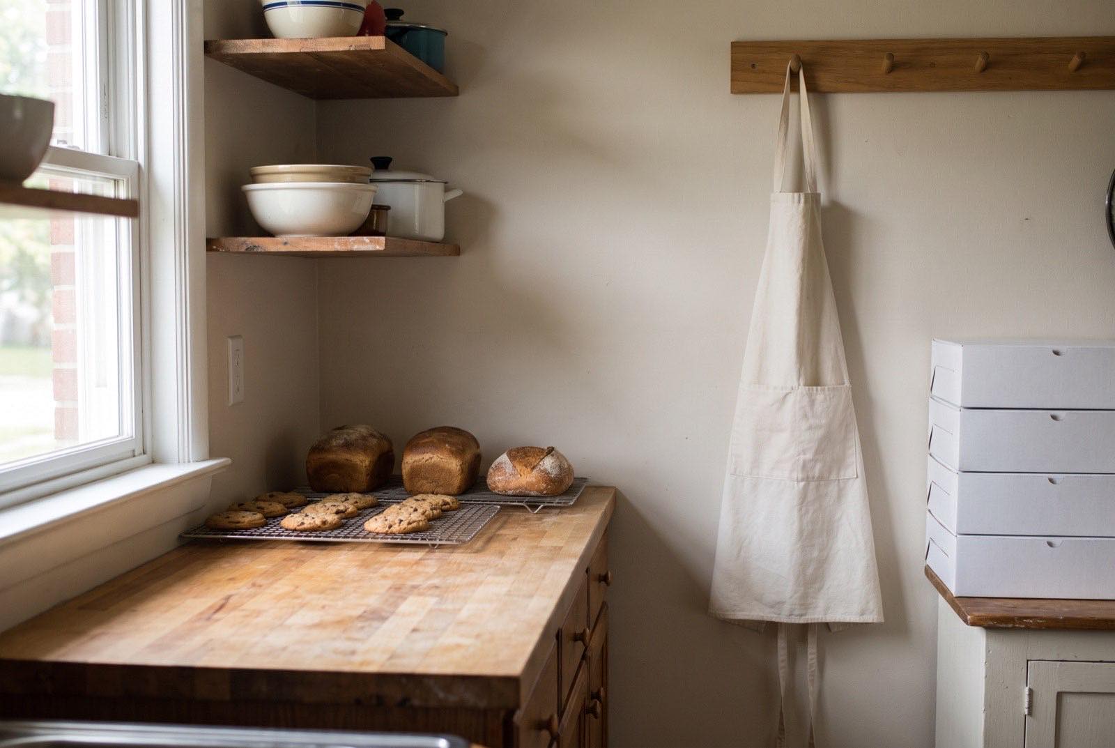 Home kitchen corner with loaves and cookies cooling, an apron on a peg rail and a stack of plain white boxes