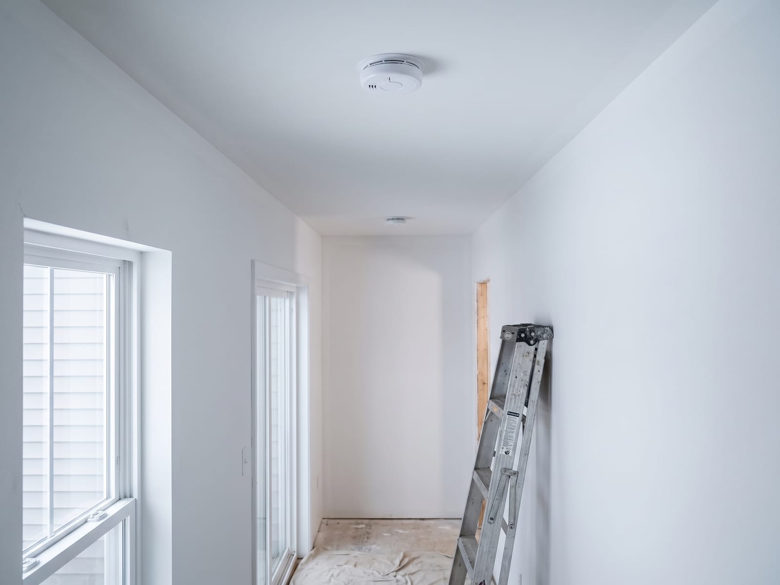 Hardwired smoke alarms centered on a freshly painted hallway ceiling, a stepladder against the wall