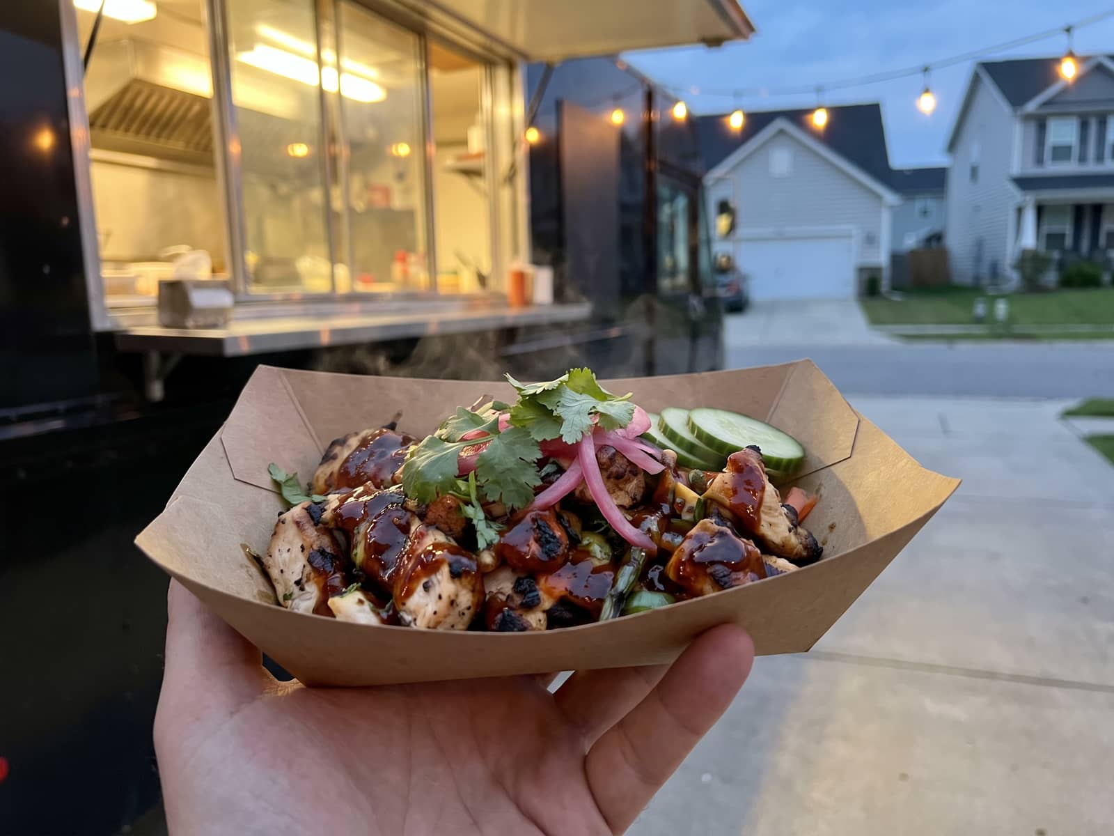 Loaded street-food portion in a plain kraft tray, charred protein with fresh herb and pickle