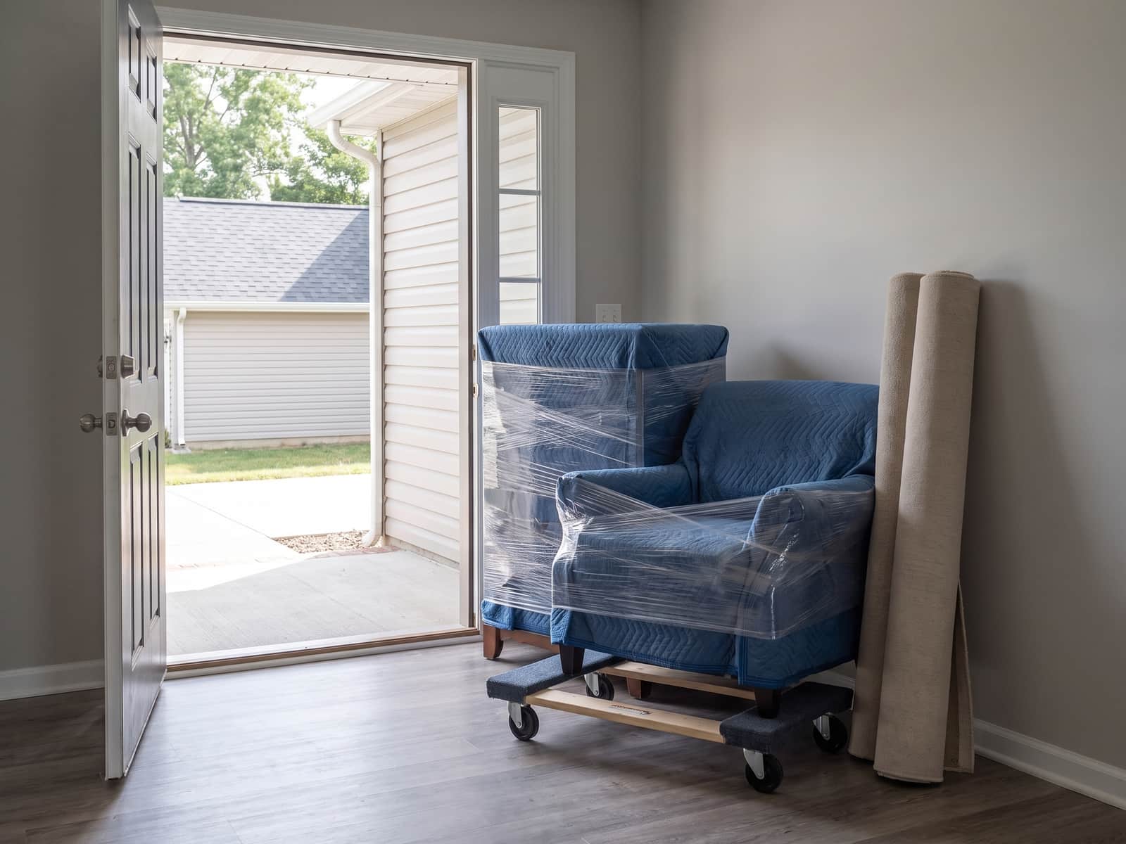 A dresser and armchair wrapped in blue moving blankets on a furniture dolly by an open front door, rolled rugs beside them