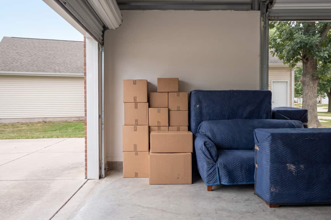 Storage space part-loaded with stacked boxes and a blanket-wrapped armchair, door open to daylight