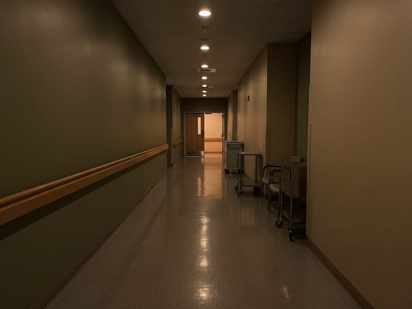 Quiet care-home corridor in the evening with a handrail and warm ceiling lights receding to an open doorway