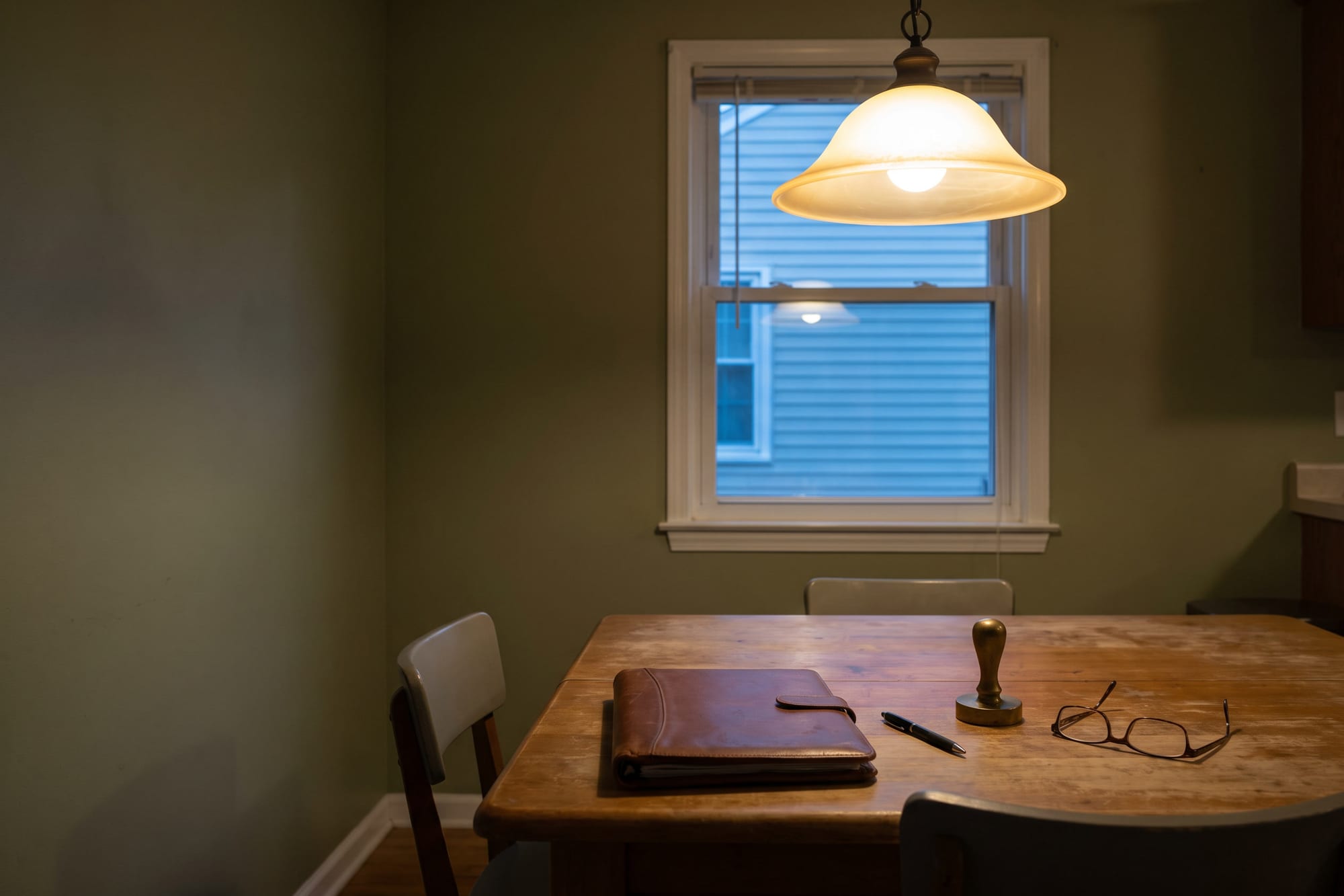 Kitchen table under a warm pendant lamp at evening with a closed leather folio, a pen and a plain hand seal set out