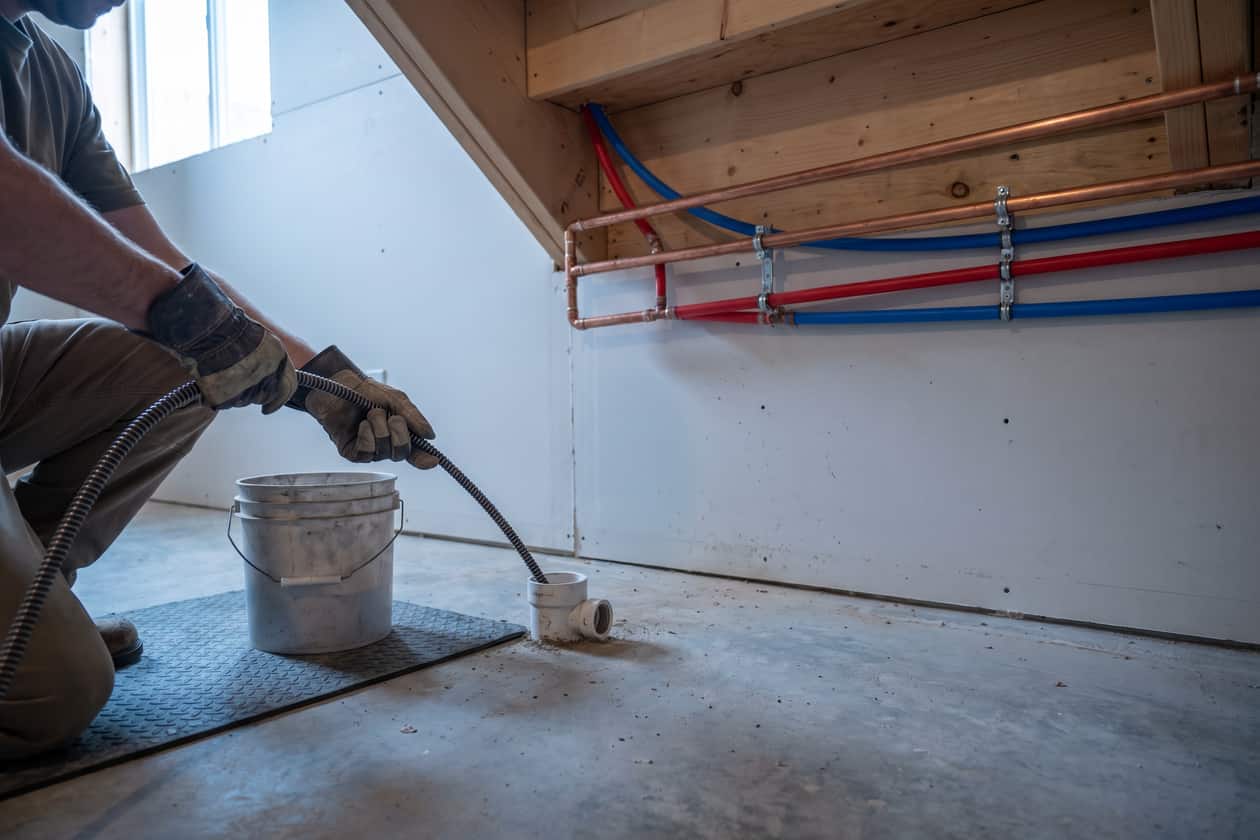 Drain auger cable fed into a cleanout with a bucket and mat protecting the floor