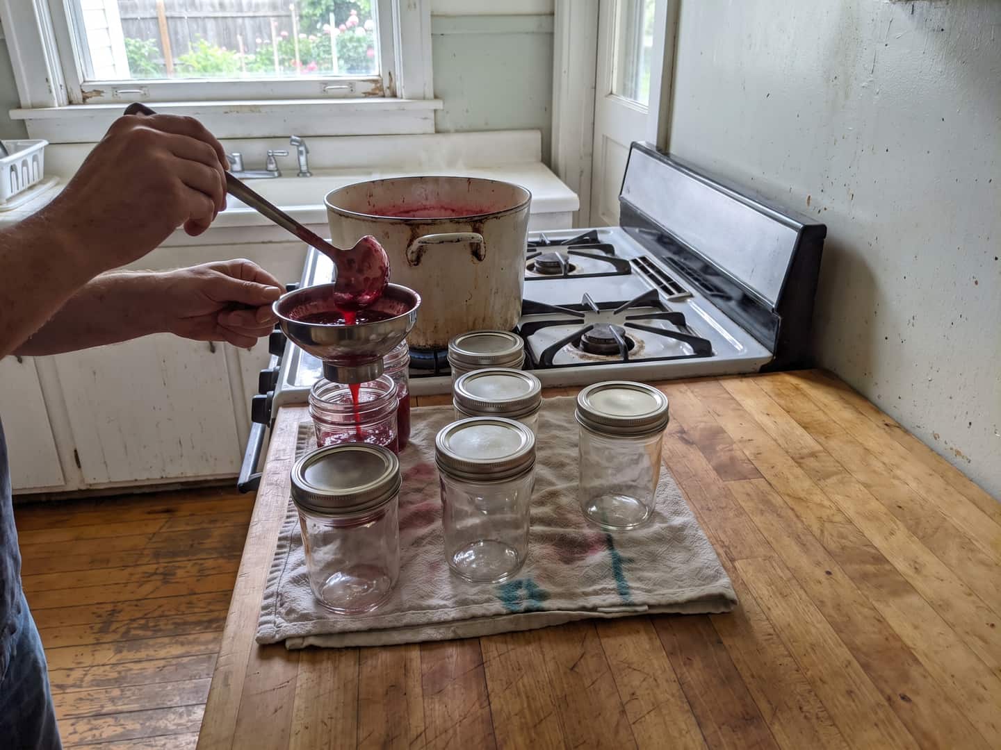 Hot jam ladled through a steel canning funnel into glass jars lined up on a folded towel on a butcher-block counter