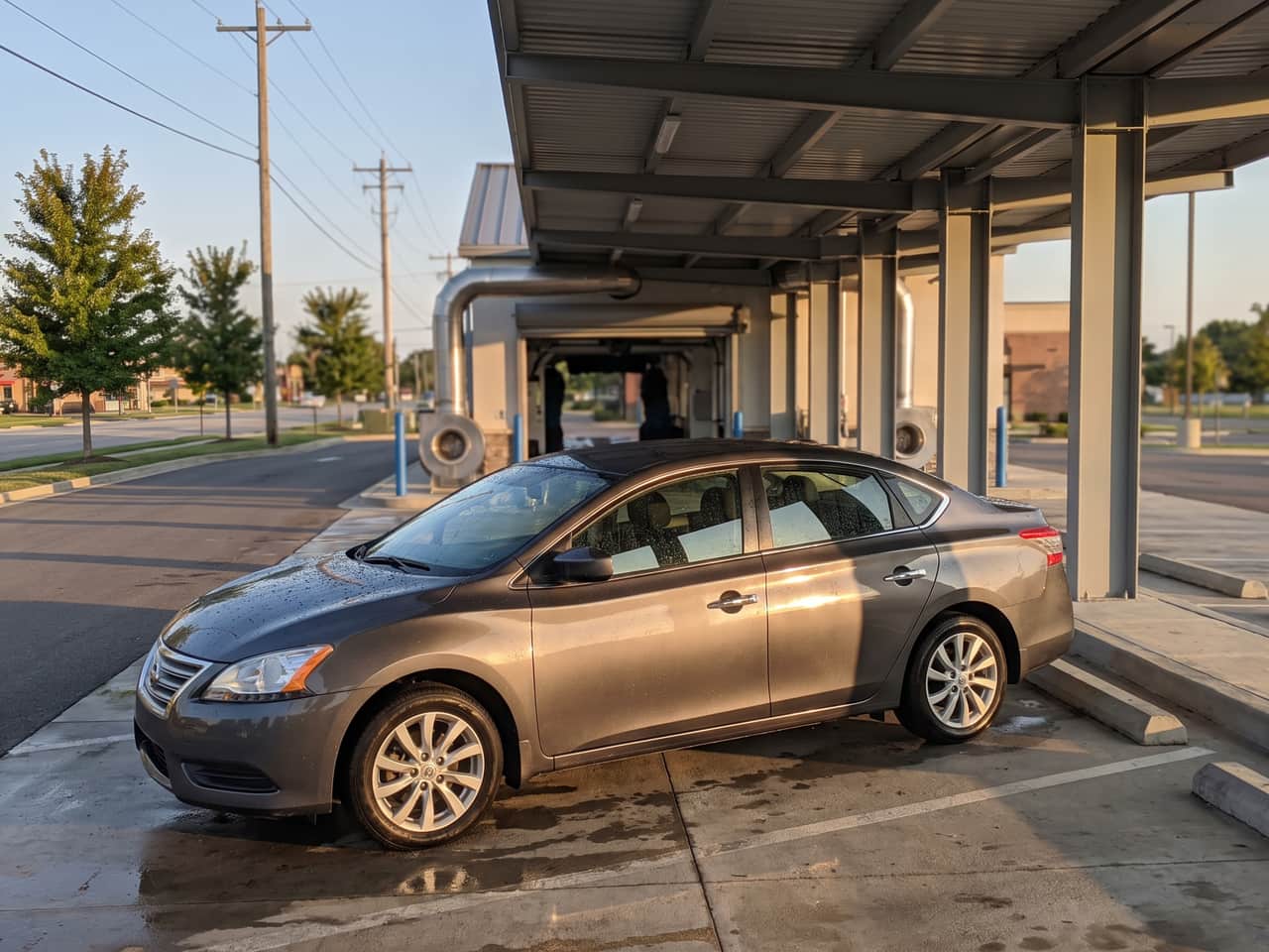Wet sedan on the concrete apron just outside the wash tunnel exit, water beaded across the hood and the drying blowers behind it
