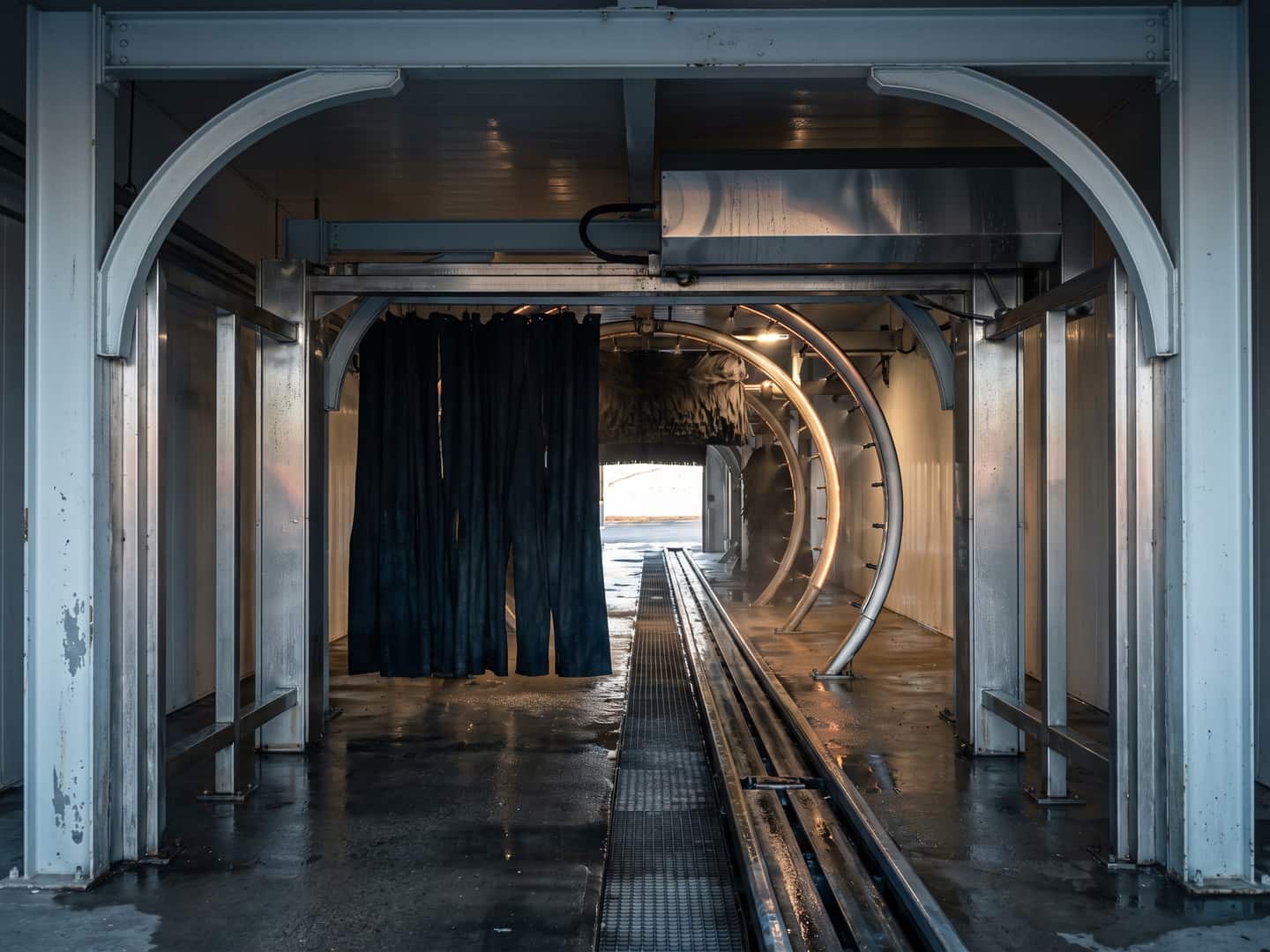 Empty wash tunnel seen down its length, conveyor track and hanging cloth curtain under the arch with daylight at the far exit
